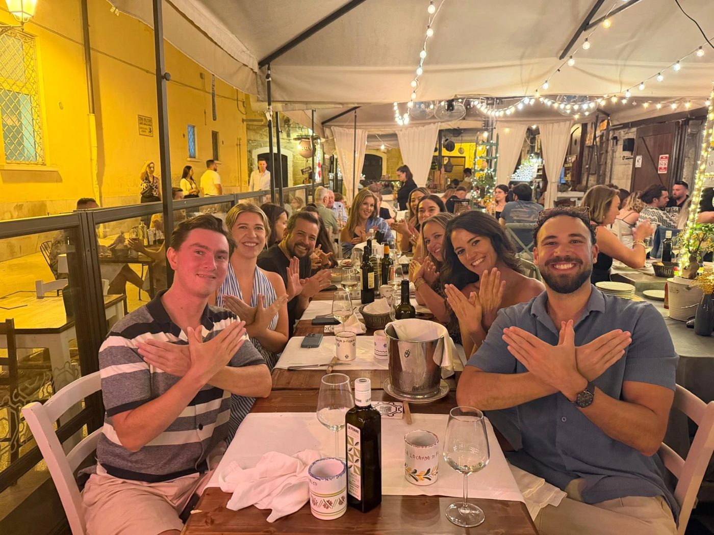 The crew at a long table under string lights on the first night in Bari, hands making the Flock sign
