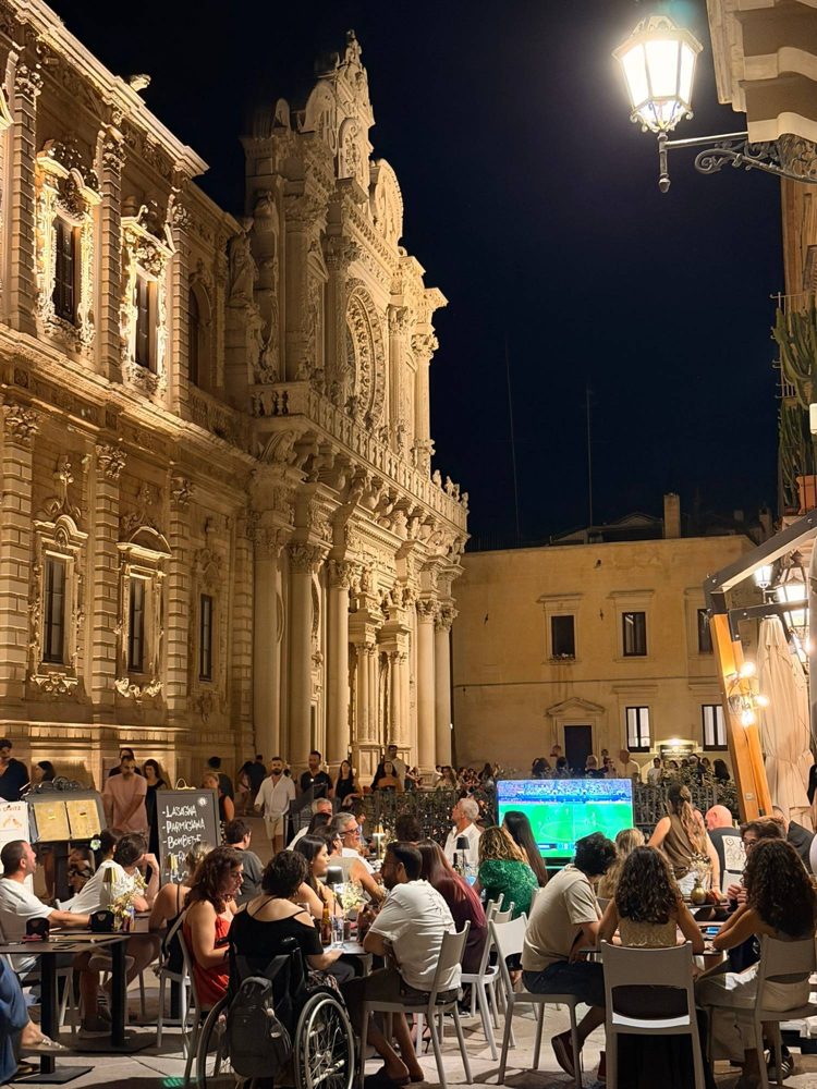 Tables filling a piazza in Lecce at night, the football on a screen