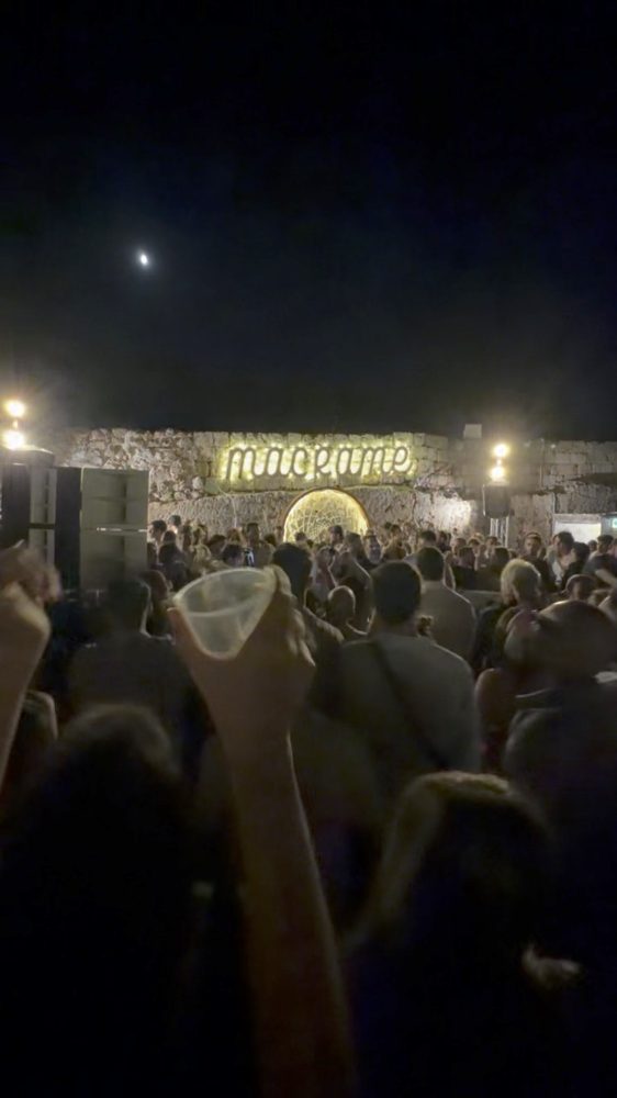 A crowd under a lit sign at a beach club at night, a cup raised