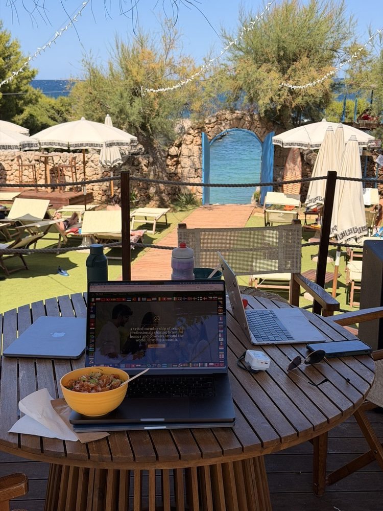 A laptop and a lunch bowl on a wooden table at a beach club, the sea through a blue gate
